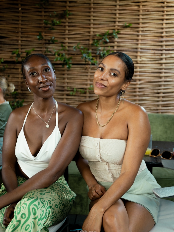 Two Women Sitting Together On An Outdoor Terrace, Dressed In Neutral Toned Summer Outfits.